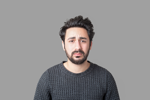 Portrait of sad young man looking at camera with frustrated facial expression against gray background. Horizontal composition. Studio shot.