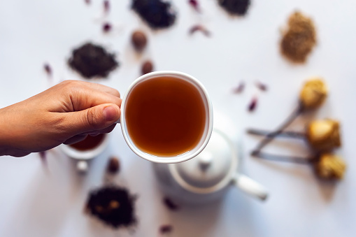 Hand holding tea cup. Woman holding a cup of tea with tea pot, dried rose flowers and other tea ingredients on the background. Flat lay view