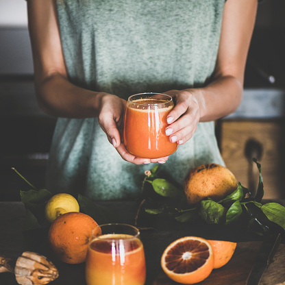 Young female holding glass of freshly squeezed blood orange juice or smoothie in hands near concrete kitchen counter, square crop. Healthy lifestyle, vegetarian, alkaline diet, spring detox concept
