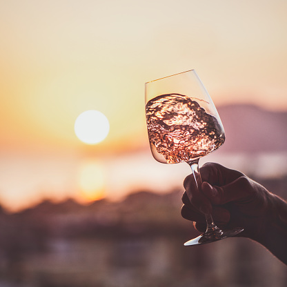 Glass of rose wine in mans hand with sea and sunset at background, close-up. Summer evening relaxed mood concept