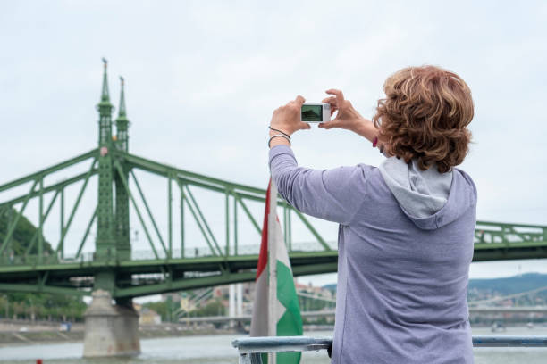 Senior woman on a deck of a cruise ship photographing a city Senior woman on a deck of a cruise ship photographing a city. budapest danube river cruise hungary stock pictures, royalty-free photos & images