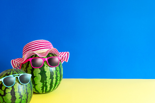 Cute summer fruits image with a boy and a girl watermelon with sunglasses and hat on a blue and yellow paper background. Cheerful summer concept.