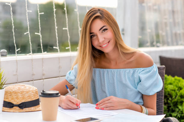 belle femme dans une robe bleue, fille en été. remplit un contrat papier, met une liste sur le contrat. cheveux longs maquillage de tous les jours. joyeux sourire. stylo à bille à la main, écrit du texte sur un morceau de papier. - paper document pen long hair photos et images de collection