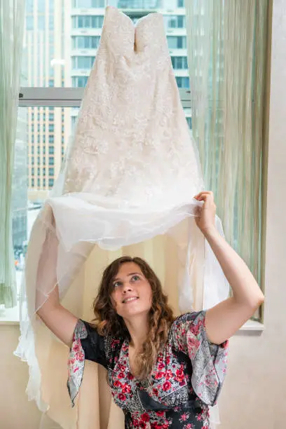 Woman young happy girl trying on wedding dress getting under by window windowsill in urban modern city hotel high rise apartment condo building before ceremony
