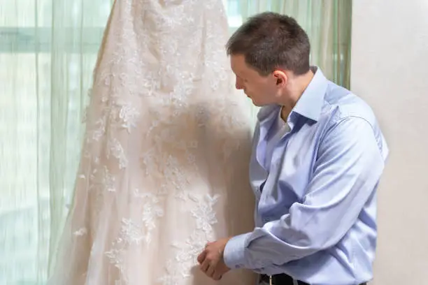 Man groom looking at wedding dress by window windowsill in urban modern city hotel apartment before ceremony