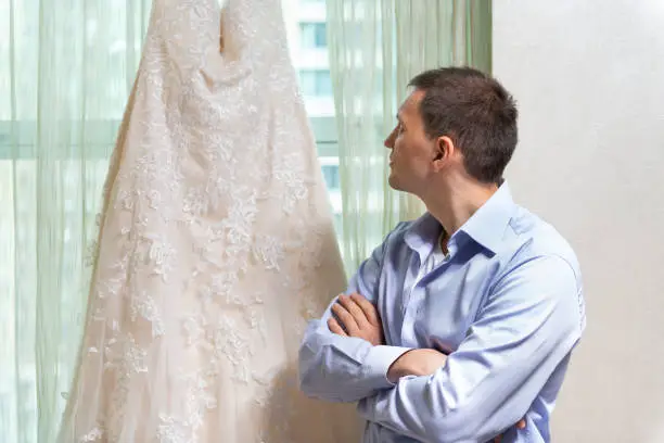 Man groom looking at wedding dress by window windowsill with arms crossed in urban modern city hotel apartment before ceremony