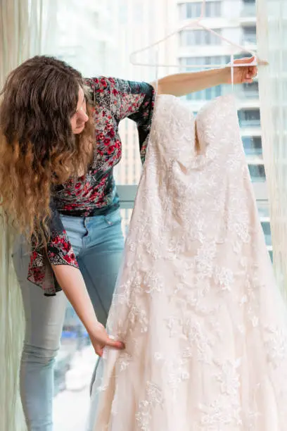 Woman young girl holding looking at wedding dress on hanger by window windowsill in urban modern city hotel high rise apartment condo building before ceremony