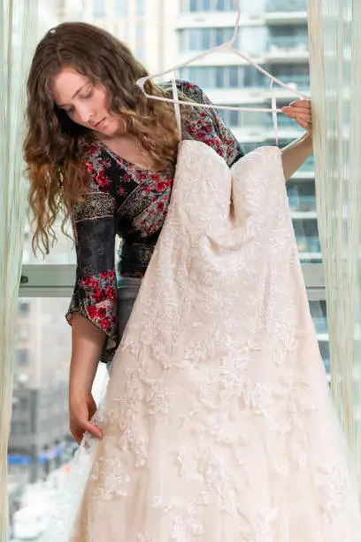 Woman young girl holding checking trying on wedding dress by window windowsill in urban modern city hotel high rise apartment condo building before ceremony