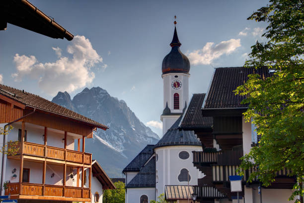 igreja st martin no centro histórico da cidade do recurso garmisch partenkirchen com o pico afiado de zugspitze de wetterstein e de nuvens brancas do montão no fundo no dia de mola ensolarado, bayern alemanha europa - mountain zugspitze mountain mountain peak scenics - fotografias e filmes do acervo