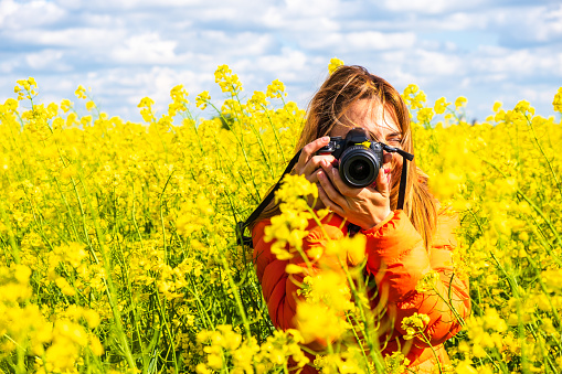 Young woman photographer with a DSLR, wearing an orange jacket, takes a picture in a rapeseed field, rural countryside, Eastern Europe, Romania - copy space on the left. Portraits, photo tours.