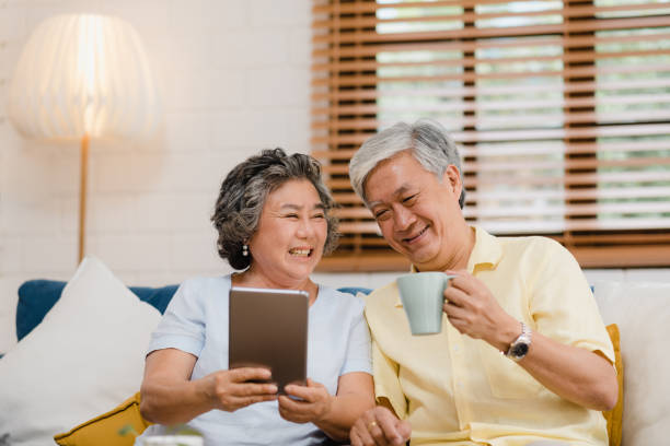 coppia di anziani asiatici che usa il tablet e beve caffè nel soggiorno di casa, la coppia si gode il momento dell'amore mentre è sdraiata sul divano quando è rilassata a casa. godersi il tempo stile di vita famiglia senior a casa concetto. - ipad senior adult facebook sofa foto e immagini stock