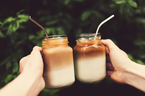 Two glass jars of ice coffee metal straws in woman and man hands. Clink cups of coffee.  Outdoor shot. Lifestyle, picnic vibes. Trendy toning. Copy space for your text.