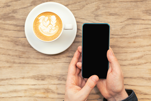 A man is holding a cellphone in hands and a cup of coffee. Flat lay, top view. Copy space for your text.