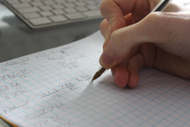 Image of college school boy doing maths homework with biro pen, teenage student studying in bedroom, writing fractions numbers and algebra formula calculations on squared graph paper, arithmetic mathematics homework, hand in front of computer keyboard Stock photo of school boy student studying and doing maths homework on paper, with close-up of the student's hand writing fractions notebook, algebra mathematics calculations by computer. math paper stock pictures, royalty-free photos & images