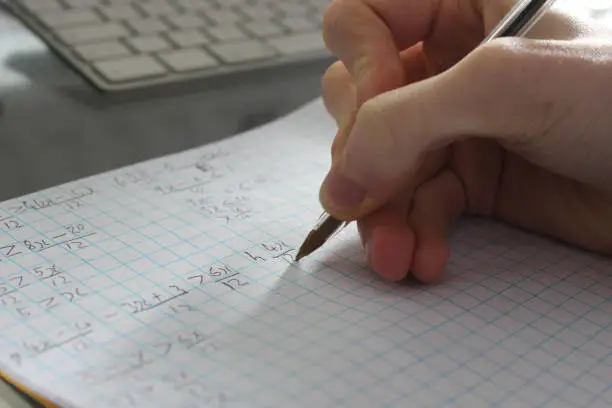 Photo of Image of college school boy doing maths homework with biro pen, teenage student studying in bedroom, writing fractions numbers and algebra formula calculations on squared graph paper, arithmetic mathematics homework, hand in front of computer keyboard