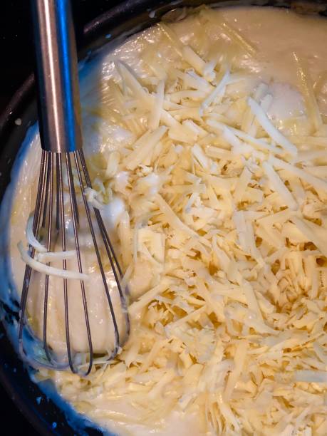 Image of cheese sauce cooking in frying pan / saucepan with bechamel white sauce (flour, butter / roux, milk) with grated Cheddar cheese, mixed with metal kitchen whisk, melting and simmering on cooker hob Stock photo of homemade cheese sauce in non-stick pan with grated mature cheddar cheese, whisking and stirring as the sauce cooks. cheese sauce stock pictures, royalty-free photos & images