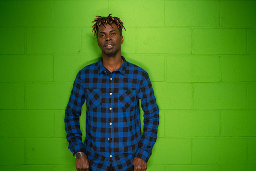 Portrait of a cool young man in front of a vivid green wall. He is wearing a blue plaid shirt.