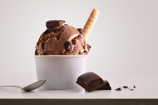 Composition of chocolate ice cream ball in paper cup on white table with products of ornament and elaboration isolated background. Horizontal composition. Front view.