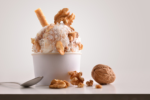 Composition of walnuts ice cream ball in paper cup on white table with products of ornament and elaboration isolated background. Horizontal composition. Front view.