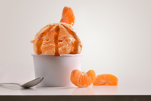 Composition of tangerine ice cream ball in paper cup on white table with products of ornament and elaboration isolated background. Horizontal composition. Front view.