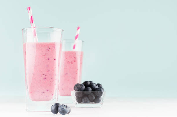 koktajle mleczne jagodowe w przezroczystym szkle z jagodami w misce, słoma w paski na stole z białego drewna i ściana w kolorze miętowym, kopiuj przestrzeń. - blueberry smoothie glass striped zdjęcia i obrazy z banku zdjęć