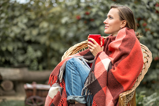 Beautiful girl resting and drinknig coffee sitting in autumn garden in a chair wrapped in a plaid woolen blanket