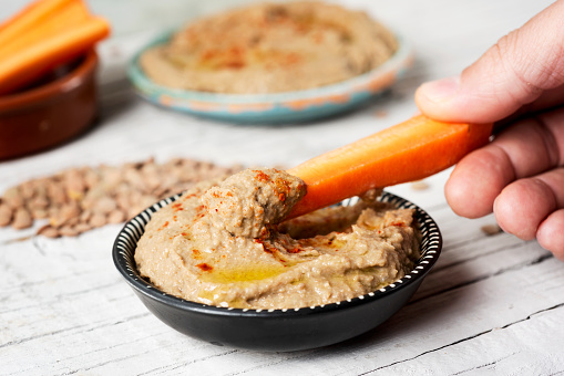 closeup of a man dipping a strip of carrot in a homemade lentil hummus seasoned with paprika served in a black and white plate, on a white rustic wooden table