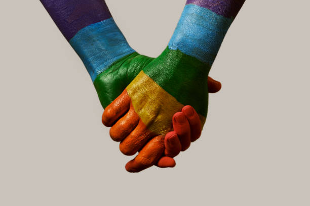 hands patterned with the rainbow flag closeup of two men holding hands, painted as the rainbow flag, against an off-white background gay pride symbol photos stock pictures, royalty-free photos & images