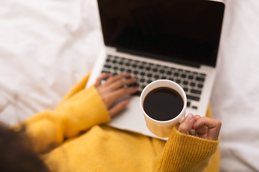 Woman surfing internet on laptop and drinking coffee in bed