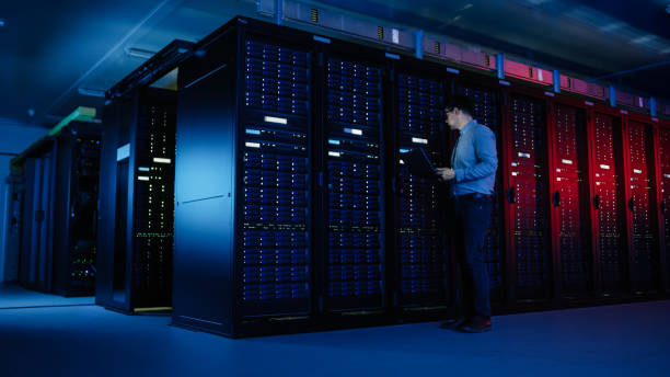 w centrum danych: male it specialist spacery wzdłuż rzędu operacyjnych stojaków serwerowych, używa laptopa do konserwacji. koncepcja telekomunikacji, przetwarzania w chmurze, sztucznej inteligencji, superkomputera - network server rack data center in a row zdjęcia i obrazy z banku zdjęć