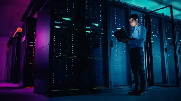 Photo of In Data Center: Male IT Technician Running Maintenance Programme on a Laptop, Controls Operational Server Rack Optimal Functioning. Modern High-Tech Operational Super Computer in Neon Colours, Lights