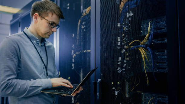 in data center it engineer stands before working server rack doing routine maintenance check and diagnostics using laptop. visible computer hardware equipment, broadband fiber optic cables led lights. - network server rack computer mainframe imagens e fotografias de stock