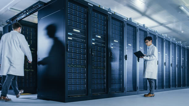 istock In Data Center: Male IT Technician Wearing White Coat Stands Beside Server Rack Cabinet, Uses Laptop Computer to Run Maintenance Diagnostics Tools. People wearing Lab Coats Working with Data. 1148195987