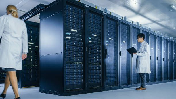 istock In Data Center: Male IT Technician Wearing White Coat Stands Beside Server Rack Cabinet, Uses Laptop Computer to Run Maintenance Diagnostics Tools. People wearing Lab Coats Working with Data. 1148196006
