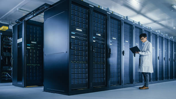 istock In Data Center: Male IT Specialist Wearing White Coat Stands Beside Server Rack, Uses Laptop Computer to Run Maintenance Diagnostics Tools, Controls so that Database Works at Optimal Level. 1148196053