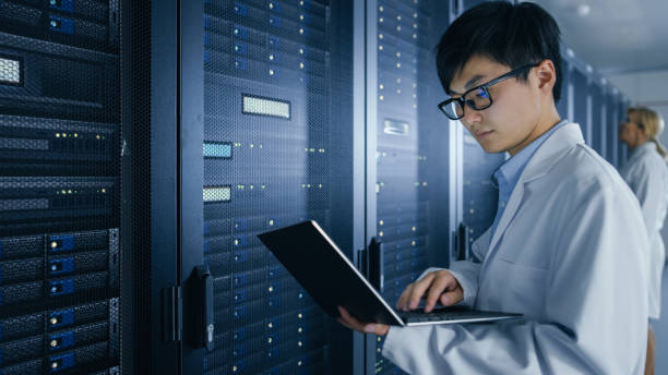 istock In Data Center: Male and Female IT Specialists Wearing White Coats Work with Server Racks, Use Laptops to Run Maintenance Diagnostics. People wearing Lab Coats Working with Datacenter Database. 1148196128