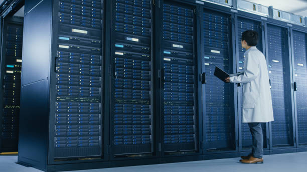 istock In Data Center: Male IT Specialist Wearing White Coat Stands Beside Server Rack, Uses Laptop Computer to Run Maintenance Diagnostics Tools, Controls so that Database Works at Optimal Level. 1148196129