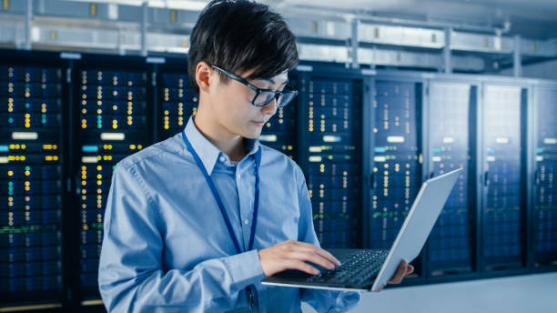 istock In the Modern Data Center: Portrait of IT Engineer Standing with Server Racks Behind Him, Doing Maintenance and Diagnostics Procedure Using Laptop. 1148196982