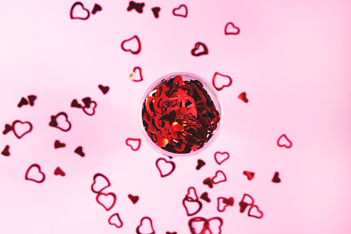 Heart-shaped vibrant red confetti in and out of champagne glass on pink background with copy space for text. Top view and close-up. Holiday, love and celebration concept.
