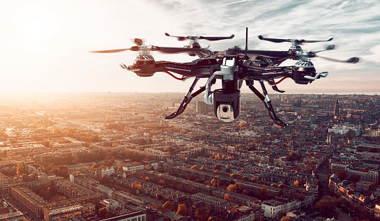 Drone flys above a city. A big camera is mounted on the drone and it can record video and take photos. It could be used for surveillance.