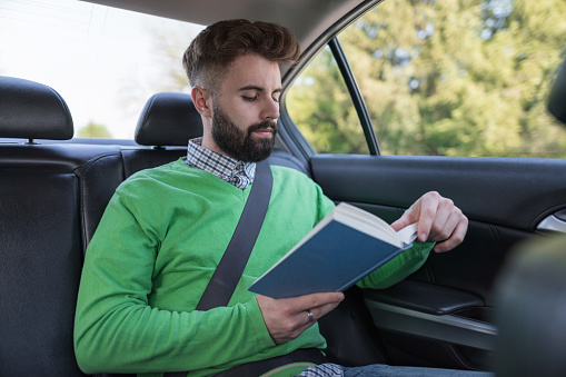 Attractive young man reading book while traveling in the car on the back seat