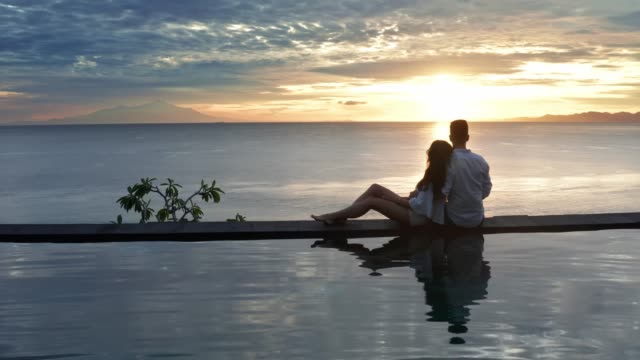 Couple In Love At Luxury Resort On Romantic Summer Vacation. People Relaxing Together In Edge Swimming Pool , Enjoying Beautiful sunset Sea View. Happy Lovers On Honeymoon Travel. Relationship, Romance. Aerial shot 4K