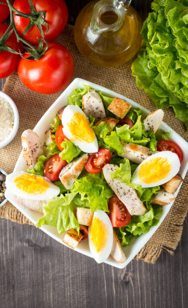 istock Fresh salad made of tomato, ruccola, chicken breast, eggs, arugula, crackers and spices. Caesar salad in a white, transparent bowl on wooden background 1148229930