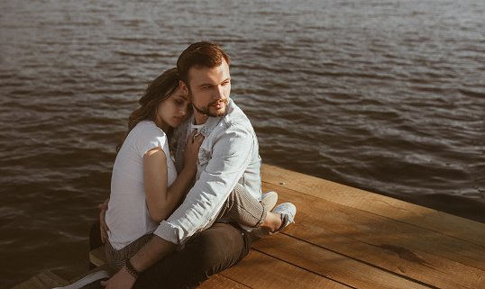 Handsome young guy looking away and hugging peacefully sleeping girlfriend while sitting on lumber pier near calm water