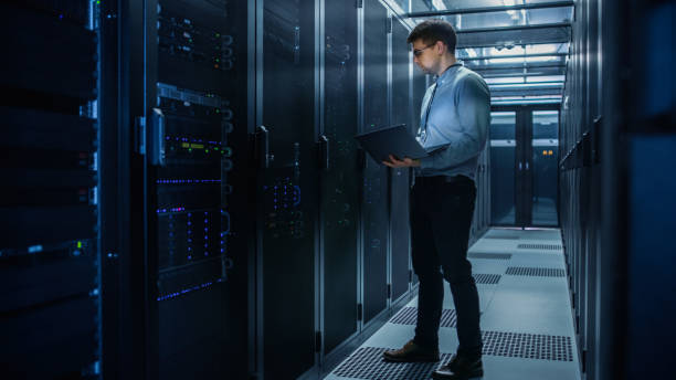 in data center it engineer stands before working server rack doing routine maintenance check and diagnostics using laptop. concept of cloud computing, artificial intelligence, supercomputer - network server rack computer mainframe imagens e fotografias de stock