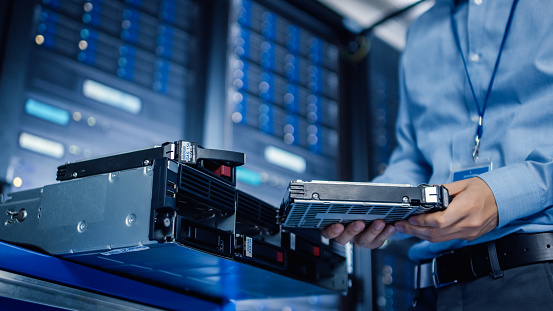 In the Modern Data Center: IT Engineer is Holding New HDD Hard Drive Prepared for Installing Hardware Equipment into Server Rack. IT Specialist Doing Maintenance and Updating Hardware.