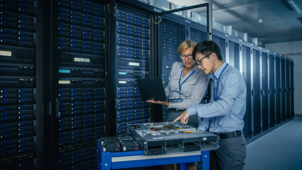 In the Modern Data Center: Engineer and IT Specialist Work with Server Racks, on a Pushcart Equipment for Installing New Hardware. Specialists Doing Maintenance and Diagnostics of the Database. In the Modern Data Center: Engineer and IT Specialist Work with Server Racks, on a Pushcart Equipment for Installing New Hardware. Specialists Doing Maintenance and Diagnostics of the Database. network server room stock pictures, royalty-free photos & images