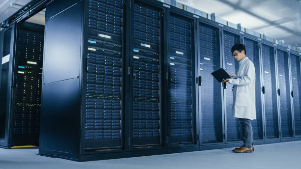 istock In Data Center: Male IT Specialist Wearing White Coat Stands Beside Server Rack, Uses Laptop Computer to Run Maintenance Diagnostics Tools, Controls so that Database Works at Optimal Level. 1148233894