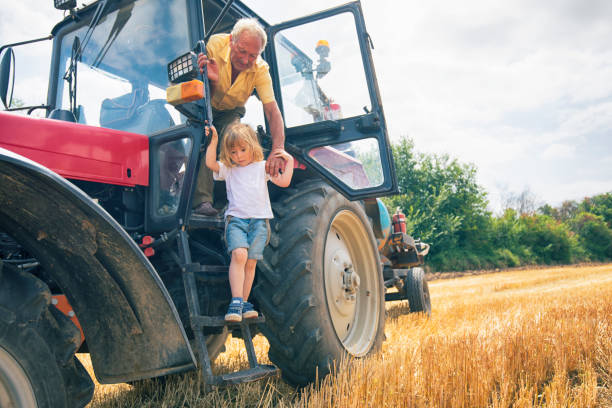 Grandfather and grandson riding a small orange tractor together in the countryside. The boy happily holds the wheel while grandpa guides him. Sunny day, bonding moment in rural setting.