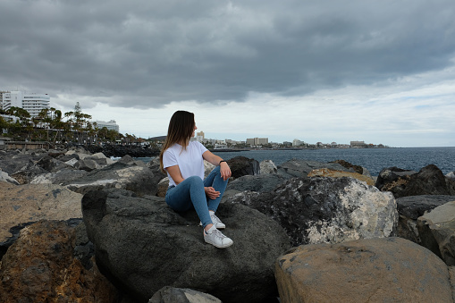 Beautiful girl sitting on beach rocks looking to the sea